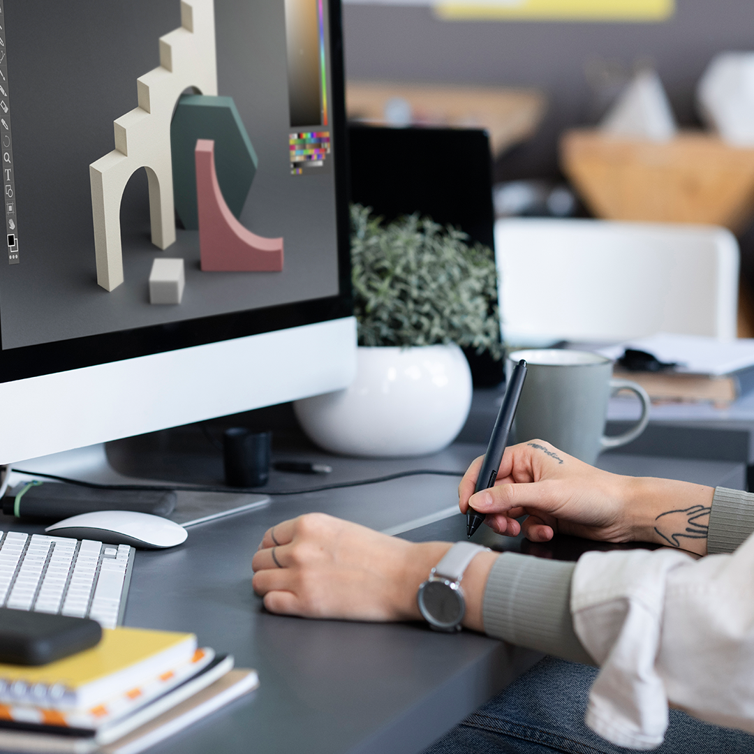 Designer working on a 3D rendering of modern shapes using a digital drawing tablet at a desk, with a computer displaying the design on the screen.