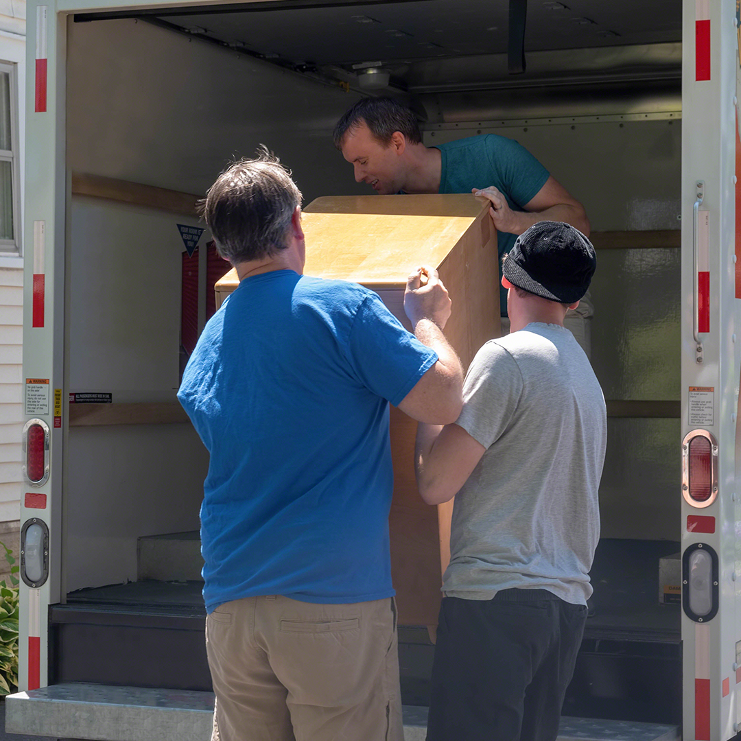 Three men carefully loading a wooden cabinet into a moving truck, teamwork and collaboration visible during the moving process.