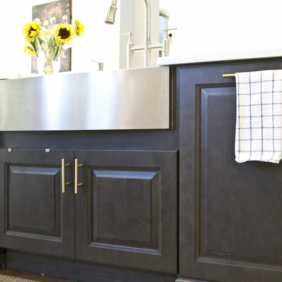 Modern dark wooden kitchen cabinets with brass handles beneath a stainless steel farmhouse sink, featuring a checkered kitchen towel and a vase of sunflowers on the countertop.