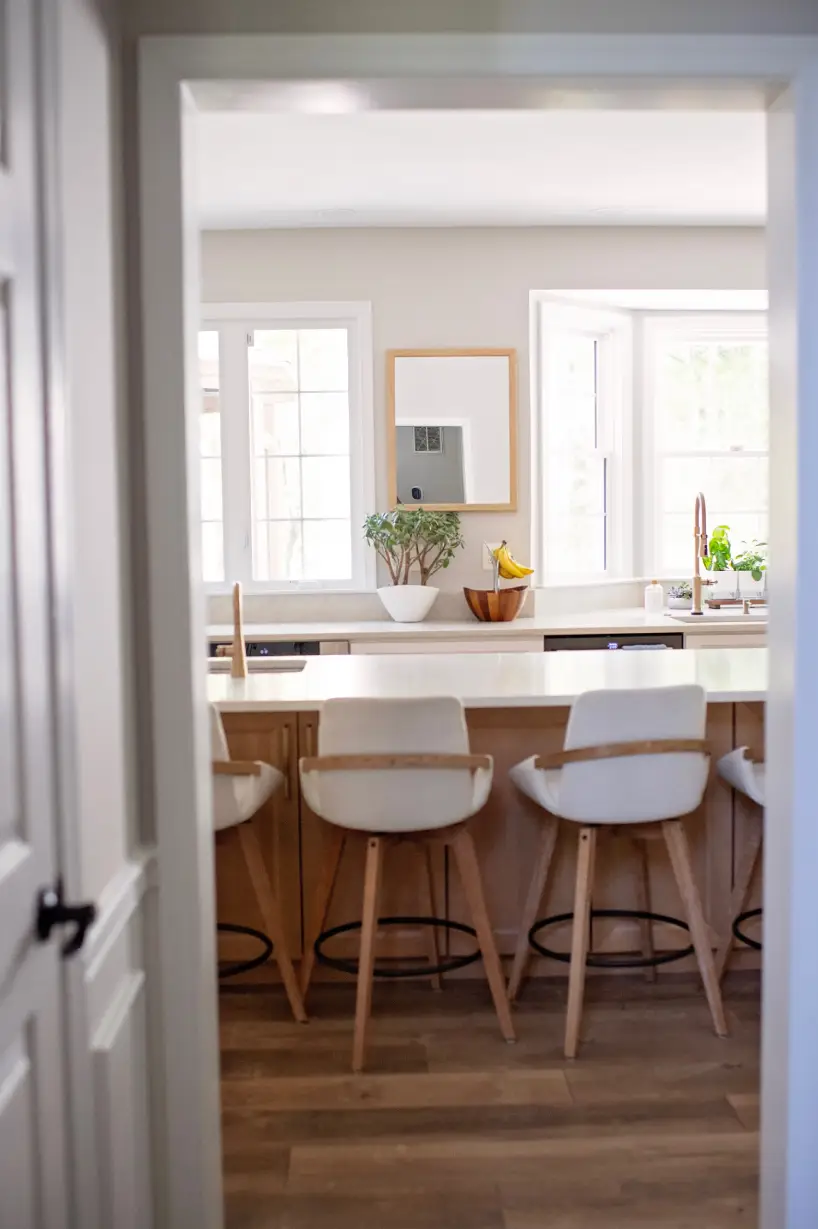Modern kitchen view through a doorway, featuring a light wood floor, white countertop with potted plant, fruit bowl, and three white barstools. Cozy and bright atmosphere.