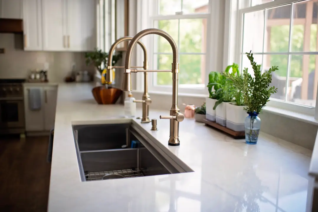 Kitchen with white countertops and dual brass faucets, set by large windows. Potted herbs line the sill, creating a fresh and airy ambiance.