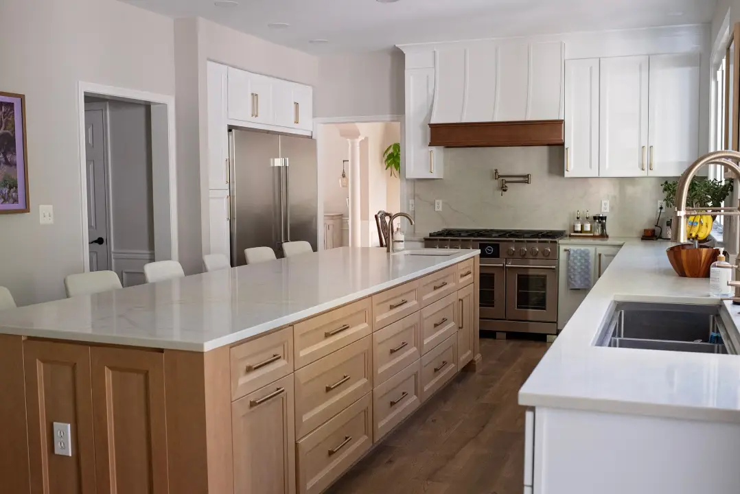 Modern kitchen with a large white island, light wood cabinets, and stainless steel appliances. Bright and inviting, with greenery by the window.