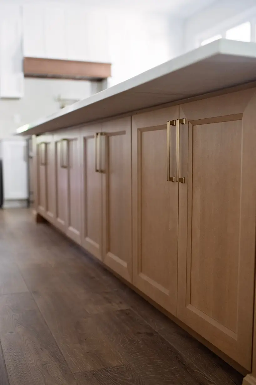 Wooden kitchen cabinets with sleek brass handles line a spacious, modern interior. The warm tones convey a cozy, inviting atmosphere.